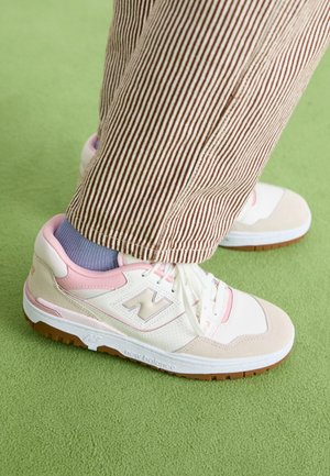 Osoba nosząca brązowe i białe spodnie w paski, jasnofioletowe skarpetki oraz beżowo-różowe buty sportowe New Balance na zielonym dywanie.