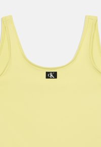 Débardeur jaune en tissu doux, avec un col rond et des bretelles. Affiche une étiquette noire avec le logo CK près du cou.