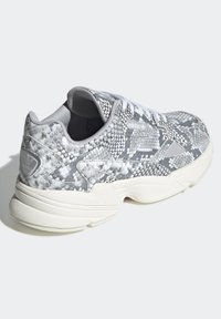 Grå sneakers med ett texturerat ormskinsmönster, vita snörning, gummisula och en Adidas-logotyp på sidan och på hälen.