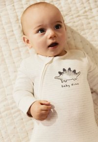 Grenouillère beige clair avec de fines rayures et un motif de stégosaure accompagné du texte "baby dino". Tissu doux avec fermeture éclair.