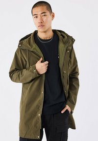 Olijfgroene waterdichte jas met capuchon, voorzien van een rits aan de voorkant, drukknoopsluiting en zijzakken. Gemaakt over een zwart t-shirt en donkere broek.