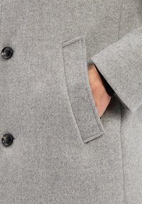 Szary płaszcz w wzór hounds-tooth o subtelnej fakturze, z czarnymi guzikami i boczną kieszenią, w której spoczywa ręka.