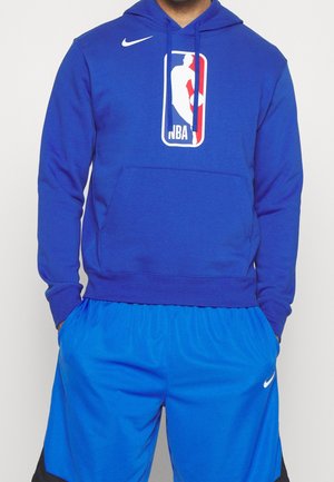 Mężczyzna w niebieskiej bluzie NBA z kapturem i dopasowanych niebieskich spodenkach do koszykówki Nike, z rękami w kieszeniach, widoczny od szyi do połowy uda.