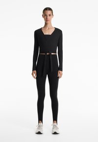 Woman standing wearing black wrap-style long sleeve top, matching high-waisted leggings, and white sneakers against white background.