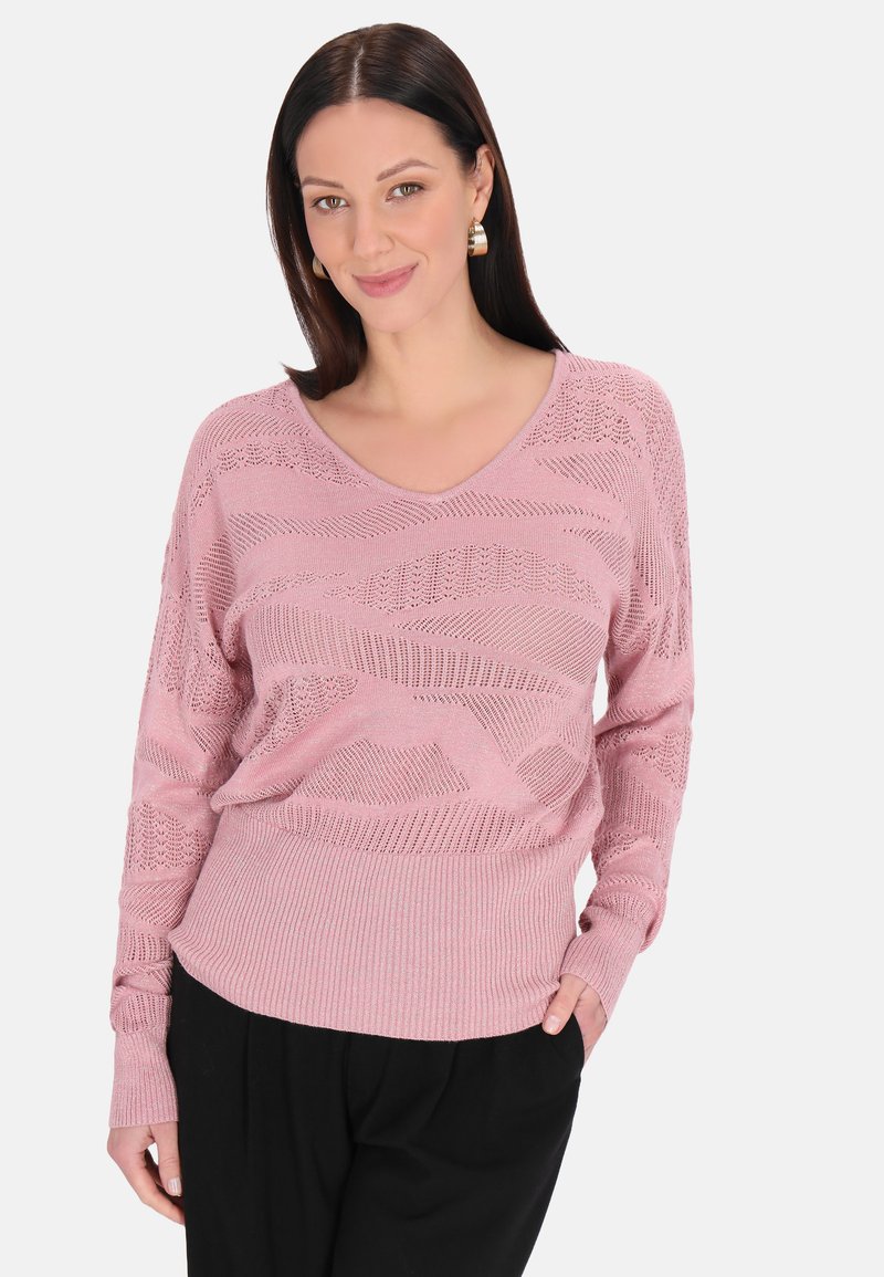 Pulover tricotat de culoare roz deschis, cu decolteu în V, modele texturate și manșete și tiv cu dungi, purtat peste pantaloni negri.
