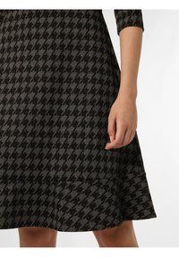 Schwarz- und grau karierte Kleid mit Dreiviertelärmeln, ausgestelltem Rock und strukturiertem Stoff. Hand auf dem Saum platziert.