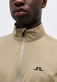 Κοντινό πλάνο ενός ατόμου που φοράει μπεζ μπουφάν με ψηλό γιακά, μπροστινό φερμουάρ και μικρό μαύρο γεωμετρικό λογότυπο στο στήθος.