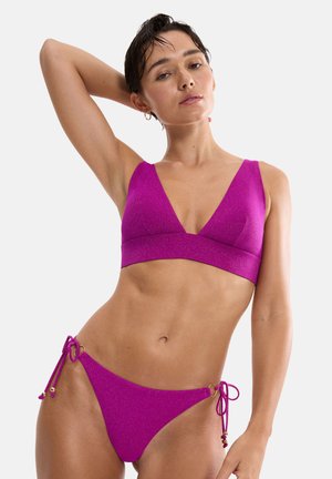 Frau mit kurzen dunklen Haaren, die einen glänzenden magentafarbenen Bikini mit seitlich gebundenem Slip trägt, posiert mit einem Arm erhoben hinter dem Kopf.