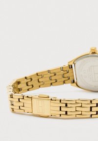 Gold metal linked watch bracelet with a clasp featuring an engraved rectangular logo on a white background.