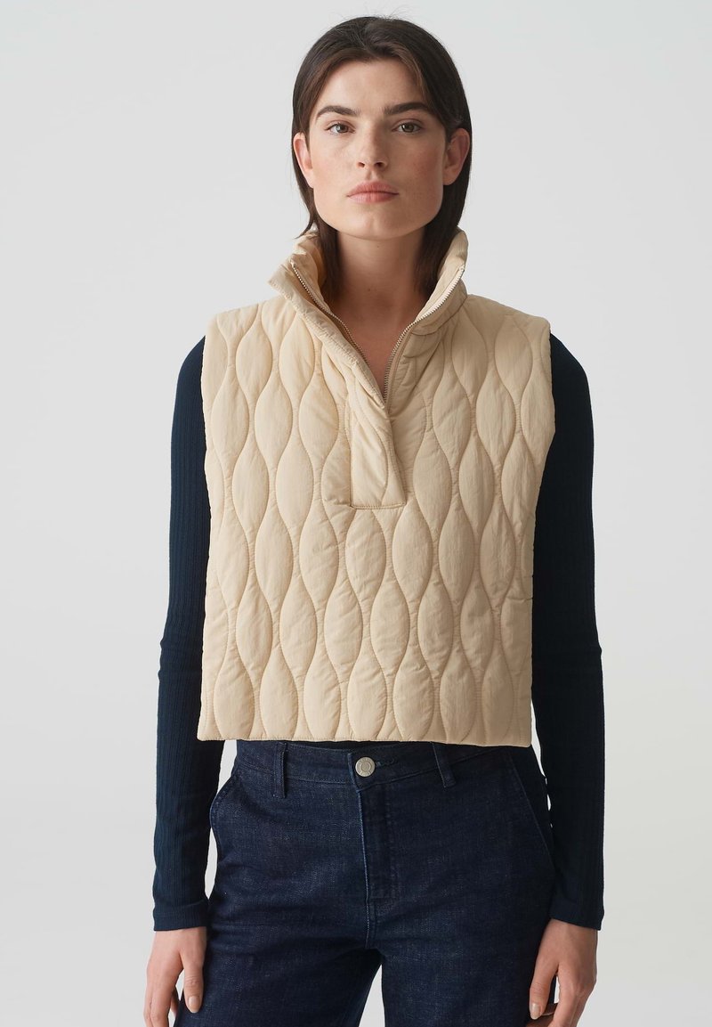 Beige gesteppte Weste mit hohem Kragen, ausgestattet mit einem halben Reißverschluss und einer aufgenähten Tasche, über einem dunklen Langarmoberteil getragen.