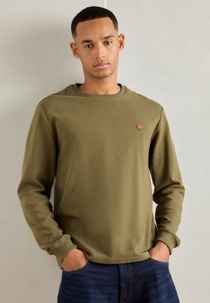 Jongeman in een olijfgroene geribbelde lange mouwen shirt met een kleine ronde patch op de borst, handen in de zakken van zijn donkerblauwe jeans, neutrale achtergrond.