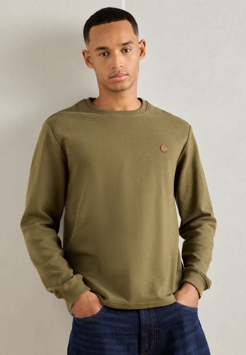 Giovane uomo che indossa una maglietta a maniche lunghe a costine verde oliva con una piccola toppa rotonda sul petto, mani nelle tasche dei jeans blu scuro, sfondo neutro.