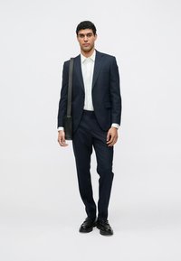 Tengerészkék öltöny szabott szabással, fehér ing és fekete cipő. Egy fekete válltáska lóg az egyik vállon. Egyszínű háttér.