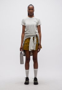 Γυναίκα που στέκεται φορώντας λευκό μπλουζάκι, μουσταρδί σορτς, γκρι πουλόβερ δεμένο στη μέση, κρατώντας γκρι παγούρι, με λευκές κάλτσες και μαύρα παπούτσια.