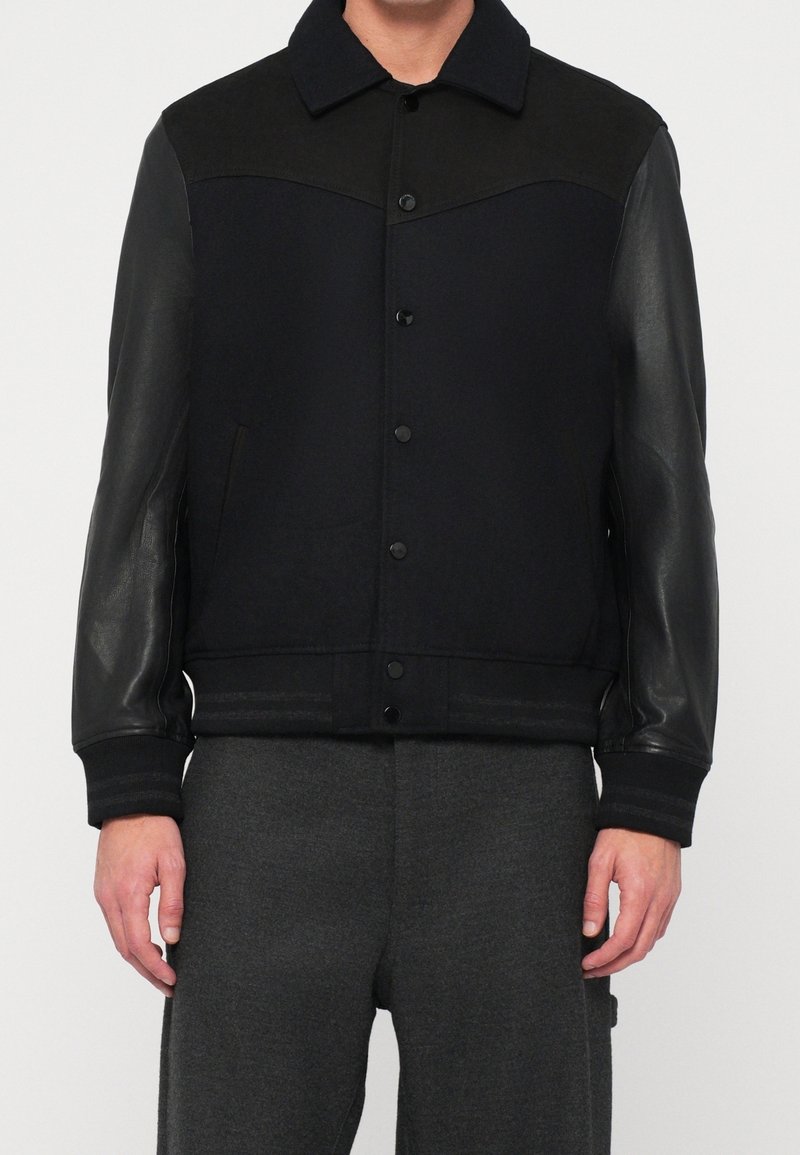Homme portant une veste noire boutonnée avec des manches en cuir et un col, associée à un pantalon gris foncé, debout devant un fond uni.