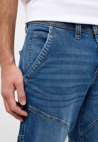 Blaue Denim-Jeans mit schmalem Schnitt, die sichtbare Nähte, Seitentaschen und einen leicht verblassten Finish für ein strukturiertes Aussehen aufweisen.