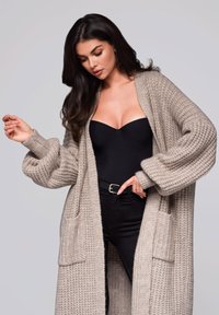 Beige gestrickte Cardigan mit lockerer Passform, geripptem Muster, zwei vorderen Taschen und weiten �Ärmeln, über einem schwarzen Body und Jeans getragen.