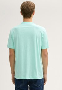 Joven de pie con la espalda frente a la cámara, vistiendo una camisa de manga corta color turquesa claro y jeans azul oscuro, contra un fondo liso.