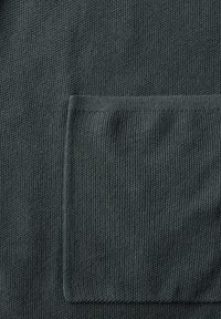 Mörkgrön texturerad tyg med en framträdande lapplomme, där ribbat kant syns längs fickan och nederkanten.