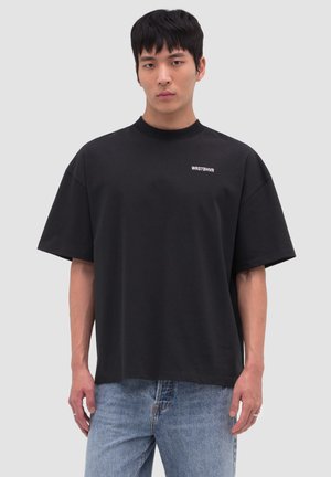 Young man stands facing forward wearing a black oversized T-shirt with "WRSTBHVR" text and light blue jeans against a plain light background.