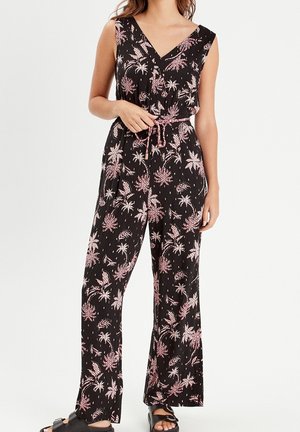 Femme portant une combinaison noire sans manches avec imprimé floral rose et ceinture à nouer, associée à des sandales noires à bout ouvert.