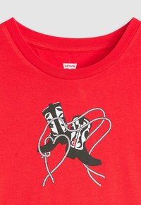 Červené tričko s grafickým designem černobílých bot propletených s bílou šňůrou. Etiketa na výstřihu nese nápis "Levi's."