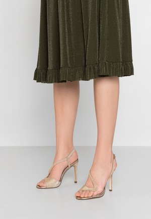 Gold strappy high-heeled sandals with a textured shimmer, open toe design, and sleek ankle straps, paired with a dark green ruffled skirt.