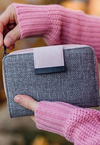 Gray woven fabric wallet with a pink zipper and strap. Features a black tag with "VUCH" on the front. Held in a hand wearing a pink knit sweater.