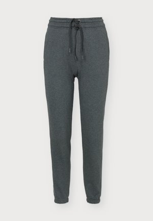 Une femme porte un haut rose sans manches, un pantalon de jogging gris foncé à cordon de serrage et des baskets blanches sur un fond clair et uni.