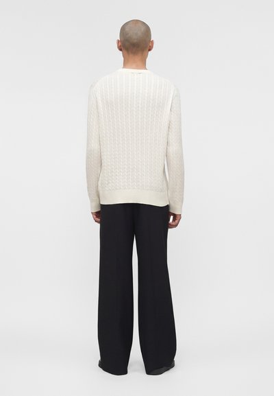 Άτομο με ξυρισμένο κεφάλι που φοράει λευκό πουλόβερ πλεκτό σε σχέδιο καλωδίων και φαρδιά μαύρη παντελόνια, στέκεται με την πλάτη προς ένα απλό φόντο.