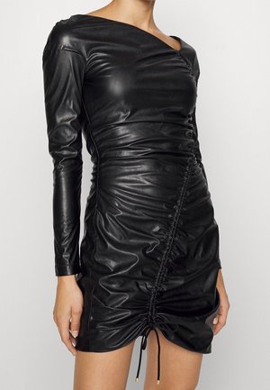 Femme portant une mini-robe moulante noire en similicuir avec des manches longues et des détails froncés sur le devant.