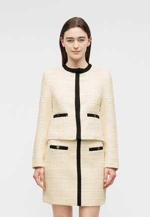 Femme aux cheveux bruns et raides portant un tailleur jupe texturé crème avec des bordures et des boutons noirs, debout devant un fond clair uni.