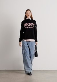 Czarny sweter z dzianiny z napisem "DKNY NEW YORK" w kolorze różowym, noszony na jasnoróżowej koszuli, w zestawie z jasnymi dżinsowymi spodniami typu cargo. Czarna torba i buty.