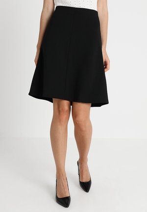 Femme portant une jupe noire en ligne A longueur genou avec des talons pointus noirs, debout sur un fond blanc uni.