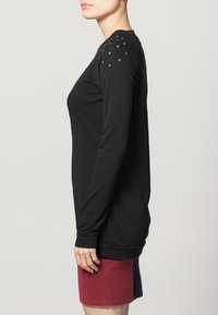 Maglietta nera a maniche lunghe con borchie decorative verdi sulle spalle, caratterizzata da un fit rilassato e polsini a costine. Indossata con una gonna multicolore.