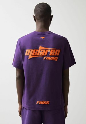 Mand iført en lilla McLaren Racing t-shirt med orange logoer, set bagfra mod en ensfarvet lys baggrund.