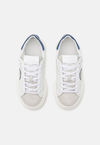 Zapatillas blancas con detalles de ante gris y una lengüeta en azul. Presentan cordones blancos y un logo en la lengüeta. Diseño de punta redondeada.