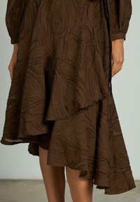 Rochie maro în straturi, realizată dintr-un material texturat cu modele intricate, tiv asimetric și margini ușor desfăcute.