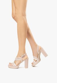 Sandali alti beige con fasce larghe, punta scoperta e chiusura con fibbia. Tacco spesso e texture liscia e lucida.