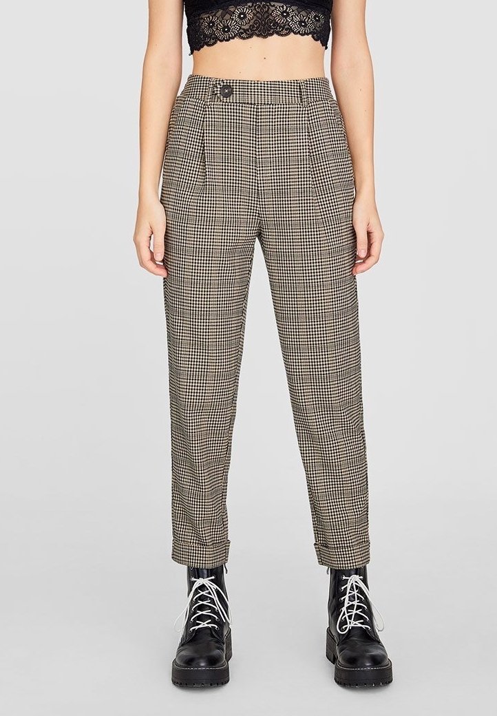 Pantalon taille haute à carreaux noir et beige, avec poches latérales et fermeture à boutons, associé à des bottes noires à lacets.