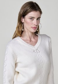 Weißer V-Ausschnitt-Pullover aus weichem Strickmaterial, mit zarter Spitzenverzierung an den Ärmeln und in der Mitte, im entspannten Schnitt.