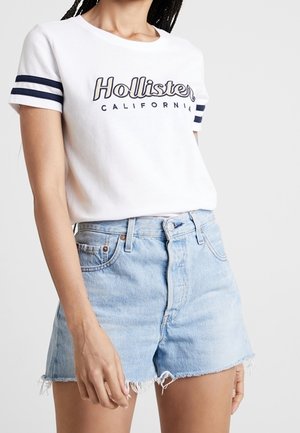 Kobieta ubrana w białą koszulkę Hollister California z granatowymi paskami na rękawach oraz jasnoniebieskie postrzępione szorty z denimu.