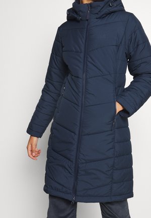 Kobieta ubrana w długi granatowy pikowany zimowy płaszcz z kapturem, zamkiem błyskawicznym z przodu oraz zapinanymi na zamek bocznymi kieszeniami, stojąca na jednolitym tle.