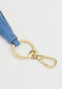 Blå lærnøkkelring med en gullmetallring og clasp. Har et duskd design med lagdelte lærstrenger. Merkegravering synlig.