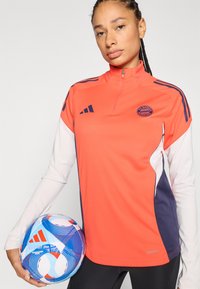 Piros és tengerészkék sportfelső negyed cipzárral, FC Bayern logóval és kontrasztos fehér ujjakkal. Egy kék-piros mintás focilabdát tart.