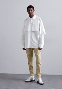 Weiße oversize Hemd mit Brusttasche, beigefarbene Hose und weiße Sneaker mit grünen Streifen. Weicher Stoff und lockere Passform.