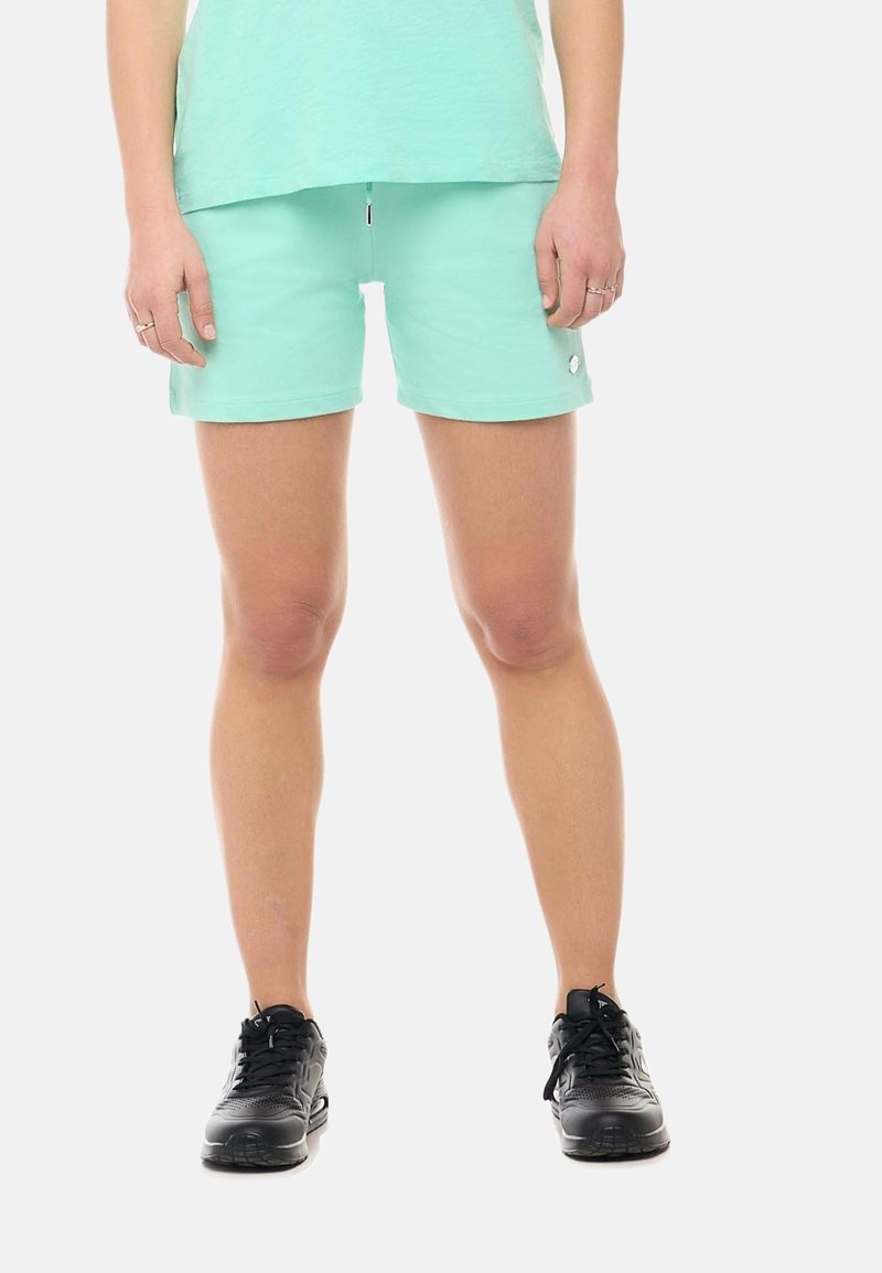 Jambes d'une personne portant un short vert clair et un haut avec des chaussures de sport noires, debout sur un fond blanc.