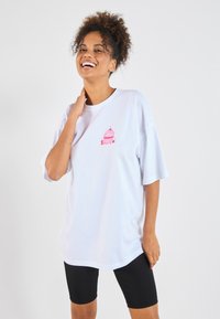 White oversized t-shirt made of cotton, featuring a pink cupcake design on the left chest area, paired with black cycling shorts.