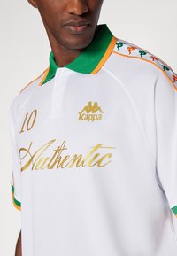 Polo blanc Kappa avec un col vert et des accents orange. L'avant comporte le texte doré "Authentique" et le numéro "10".