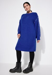 Lysblå strikket kjole med oversized silhuet, ribbede manchetter og sidesnøre detaljer, parret med sorte kraftige hæle støvler.